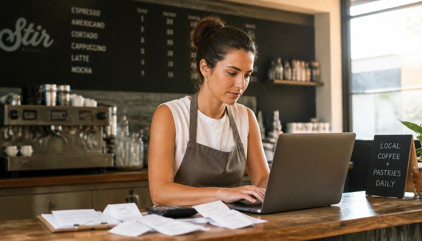 A small business owner doing bookkeeping on a laptop with receipts and a calculator at a San Diego coffee shop counter