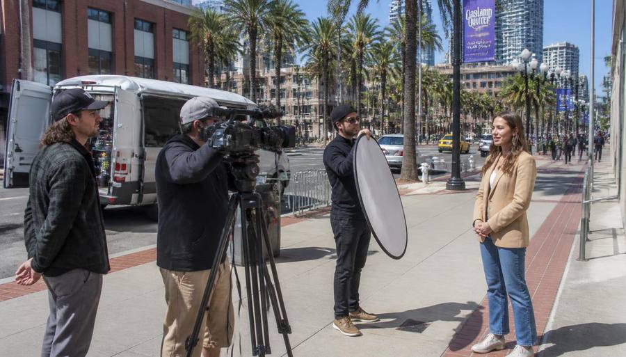A small film crew with a camera on a tripod and a reflector shooting on a palm-lined downtown San Diego street