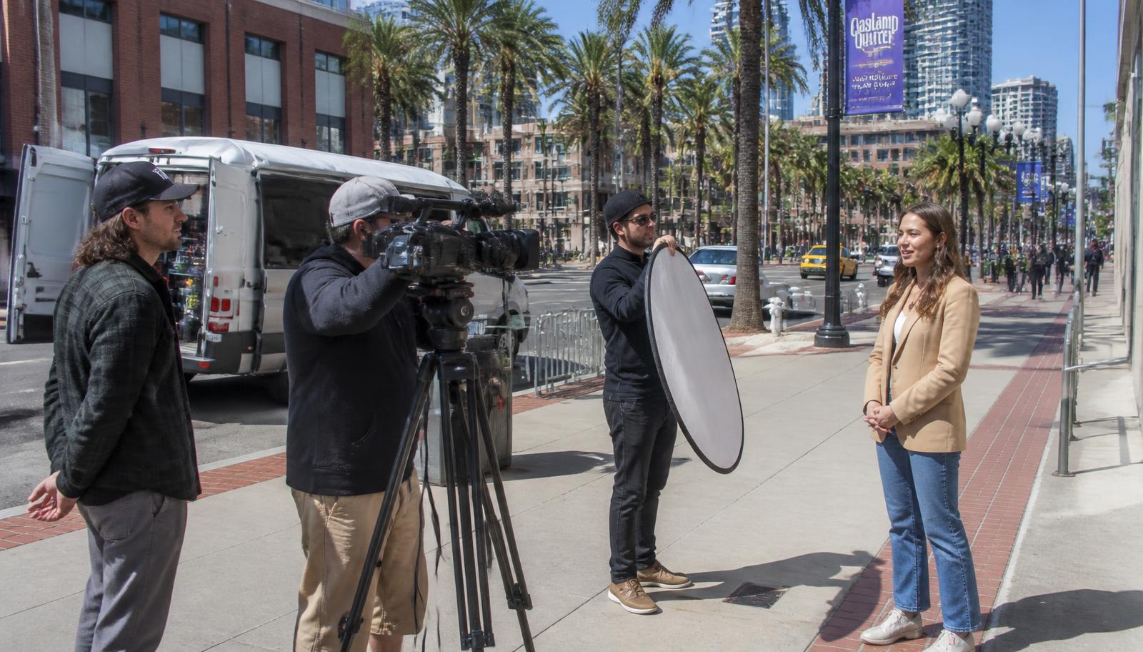 A small film crew with a camera on a tripod and a reflector shooting on a palm-lined downtown San Diego street