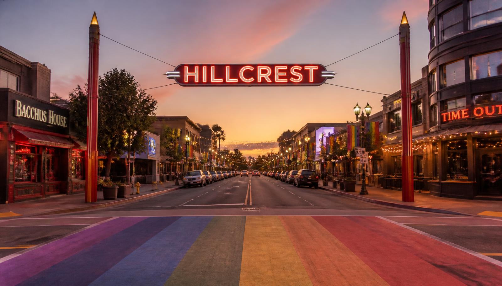 The neon Hillcrest sign glowing over University Avenue at dusk with rainbow crosswalk and lit bar patios in San Diego
