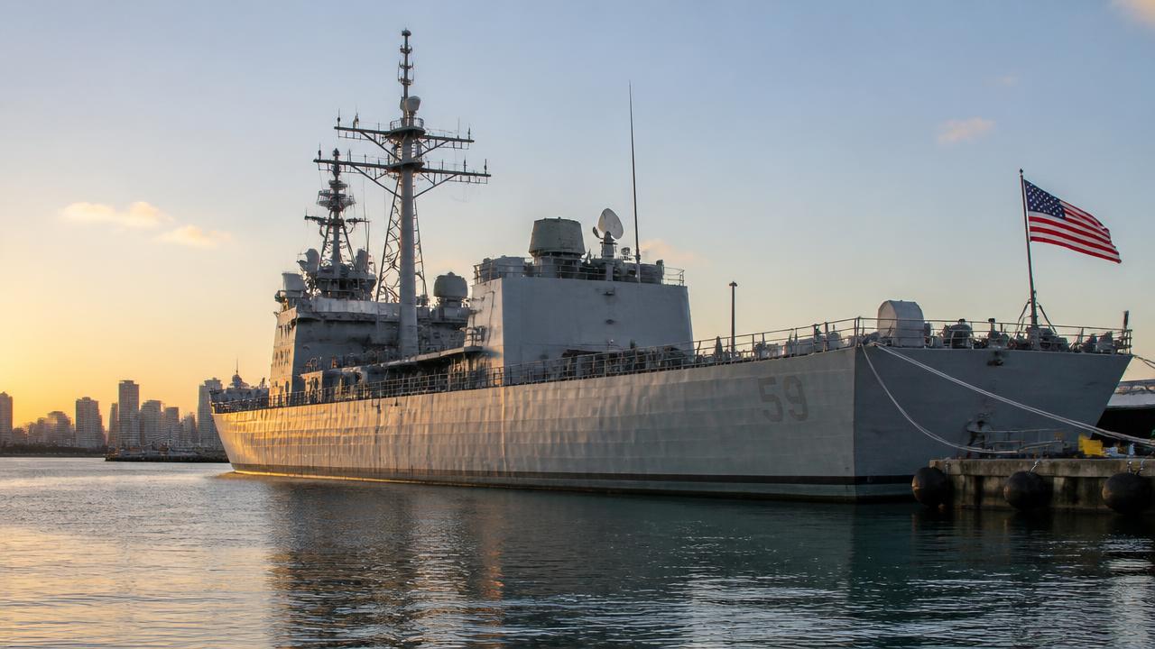 US Navy warship docked at a San Diego pier at sunrise with the American flag flying and city skyline behind