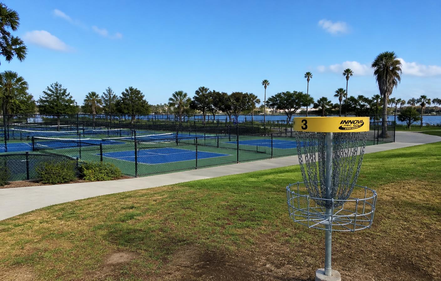 Public San Diego park with blue pickleball courts, a disc golf basket, palm trees, and a lake in the background under a clear sky